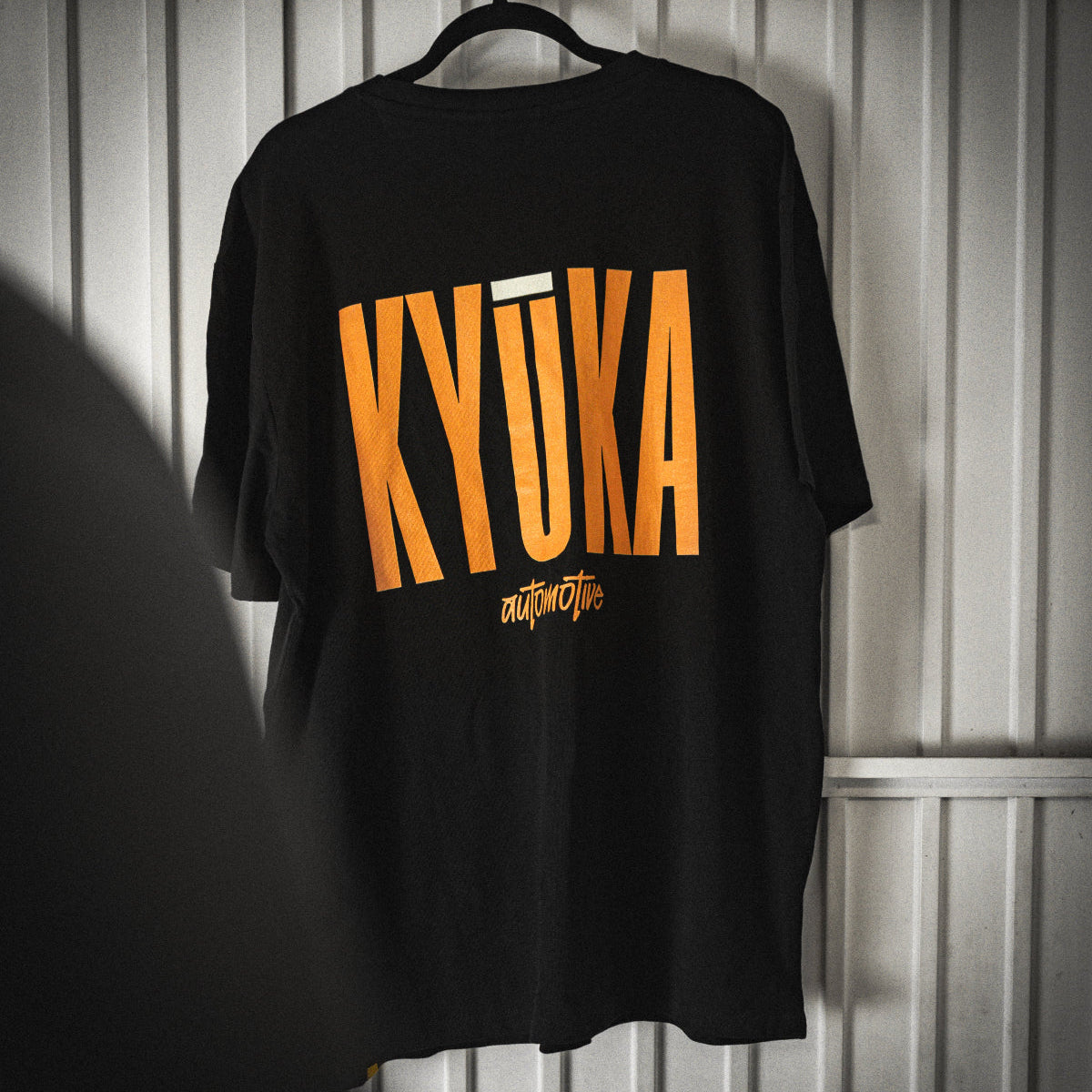 Black t-shirt with yellow 'KYUKA' text on a hanger against a metallic wall.