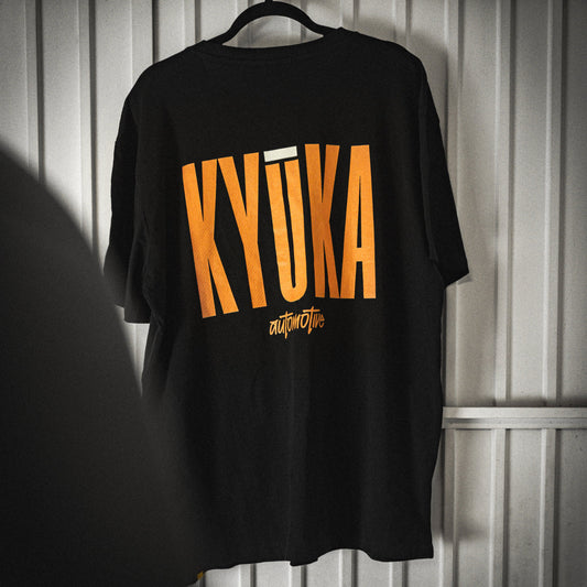 Black t-shirt with yellow 'KYUKA' text on a hanger against a metallic wall.